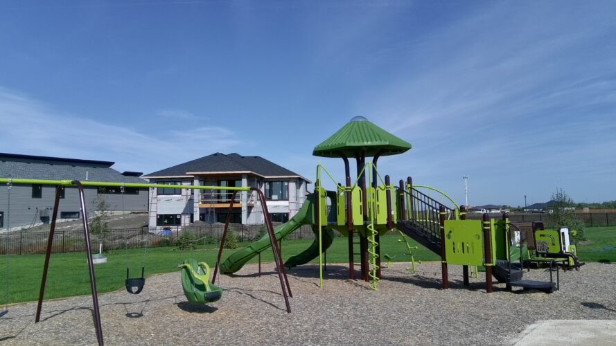 Green swings and large playground structure with homes in the background.