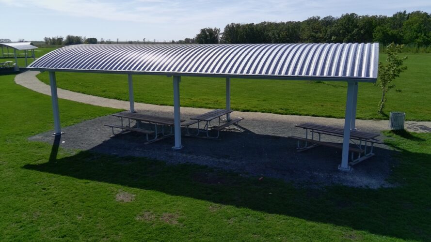 Large silver shelter providing shade to three picnic tables in a grass field.