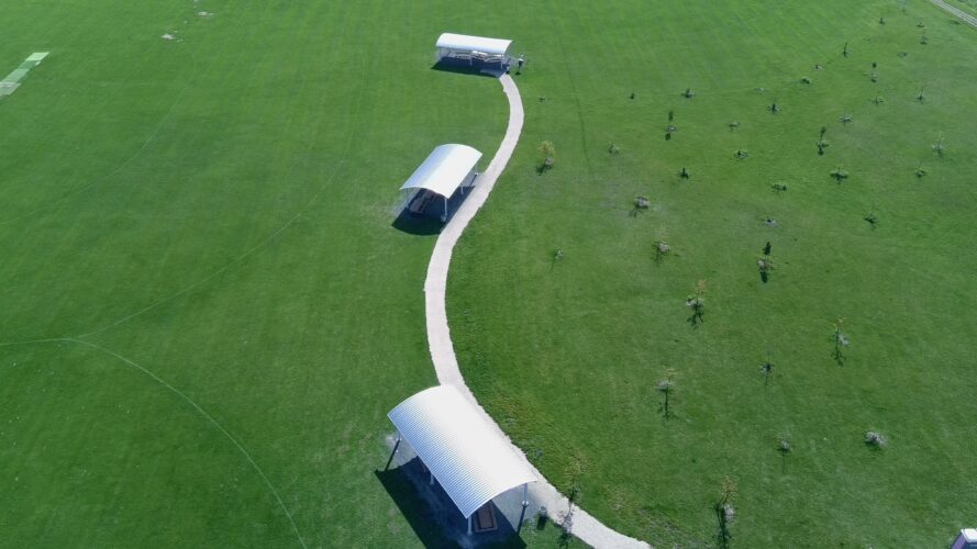 Three large commercial outdoor shelters on the path next to a cricket field.