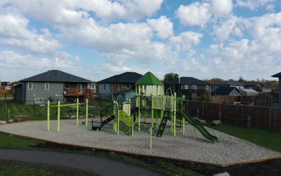 Playground structure at Amberfield Park in Manitoba, with neighborhood homes nearby.